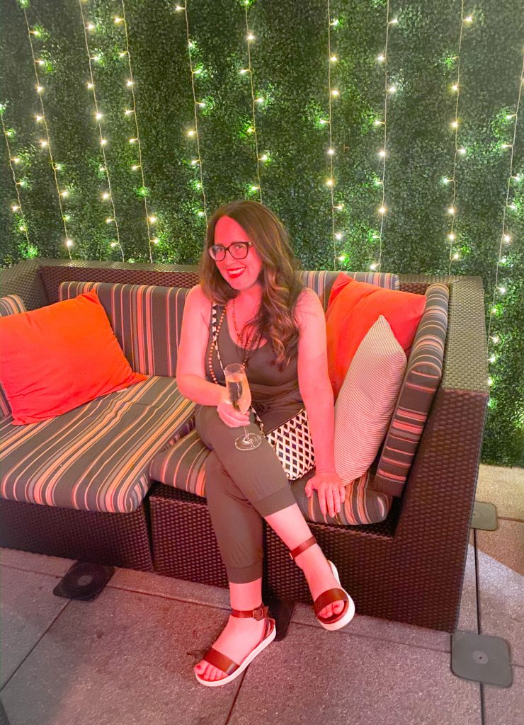 woman wearing green jumpsuit sitting on striped couch holding a glass of champagne in front of a wall of lights