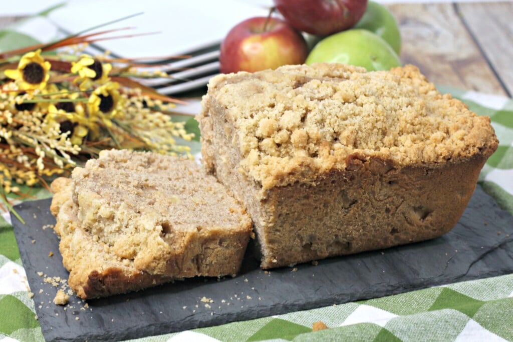 loaf of half sliced apple bread next to red and green apples, yellow sunflowers and white plates