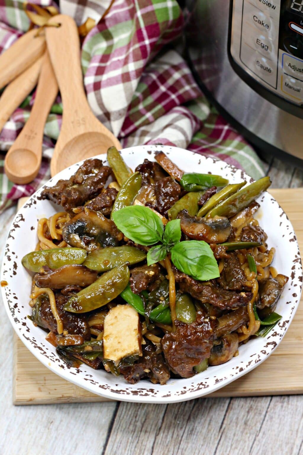 Thai steak noodles in white bowl next to wooden spoons and burgundy plaid towel
