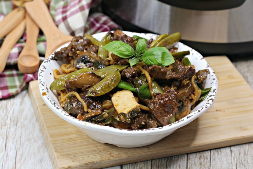 Thai steak noodle dinner in white bowl on wood tray next to wood spoon and burgundy plaid towel